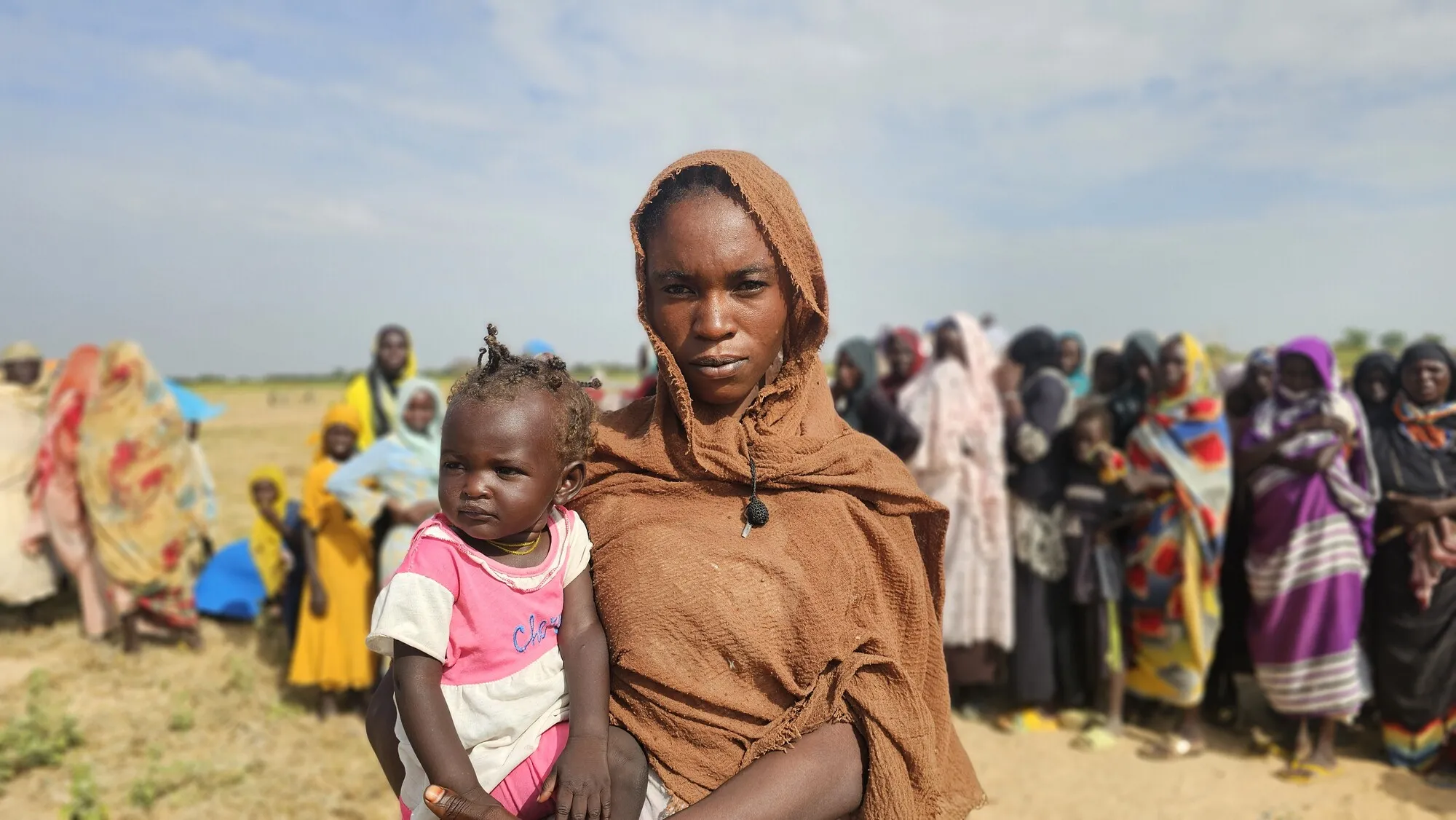 A Sudanese woman holds her baby daughter while a crowd of people stands in the background.