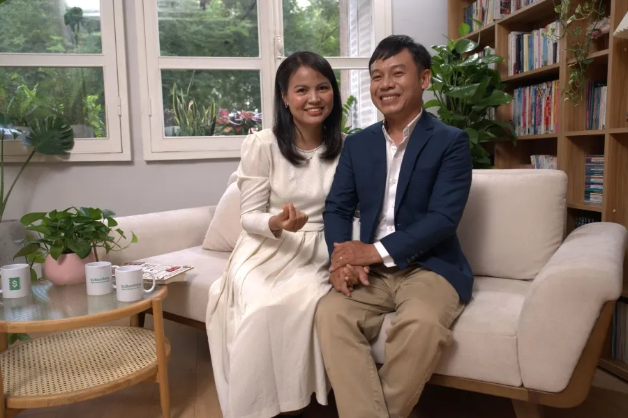 A Vietnamese couple, holding each other's hands, smiling at the camera while sitting on a sofa.