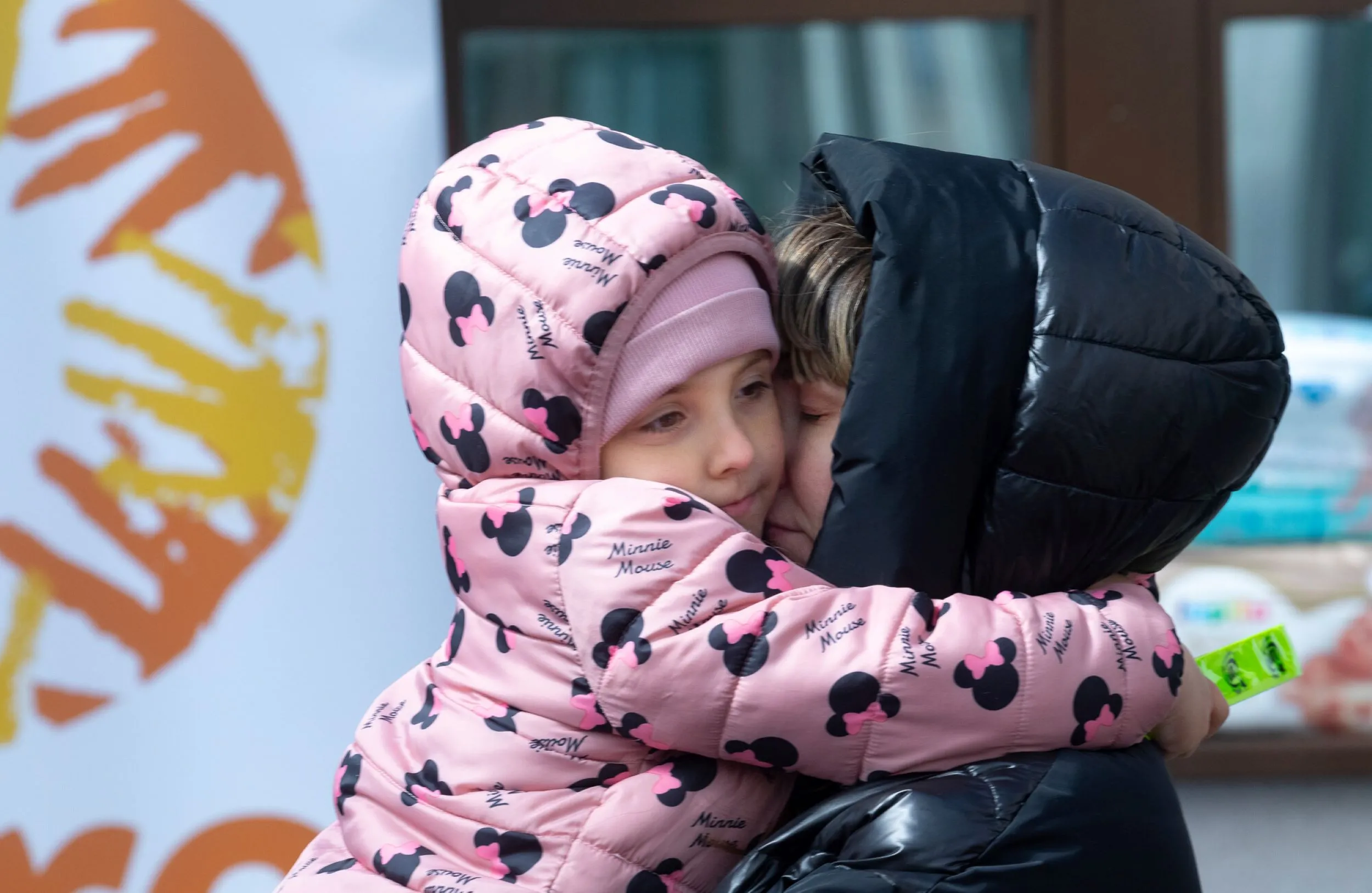 A young Ukrainian child in a pink Minnie Mouse jacket is in a close embrace with an adult, presumably her mother, who is wearing a black puffer jacket and has her eyes closed. The child looks up with a serene expression, while the mother’s face appears loving and protective. A CARE logo is partly visible in the background.