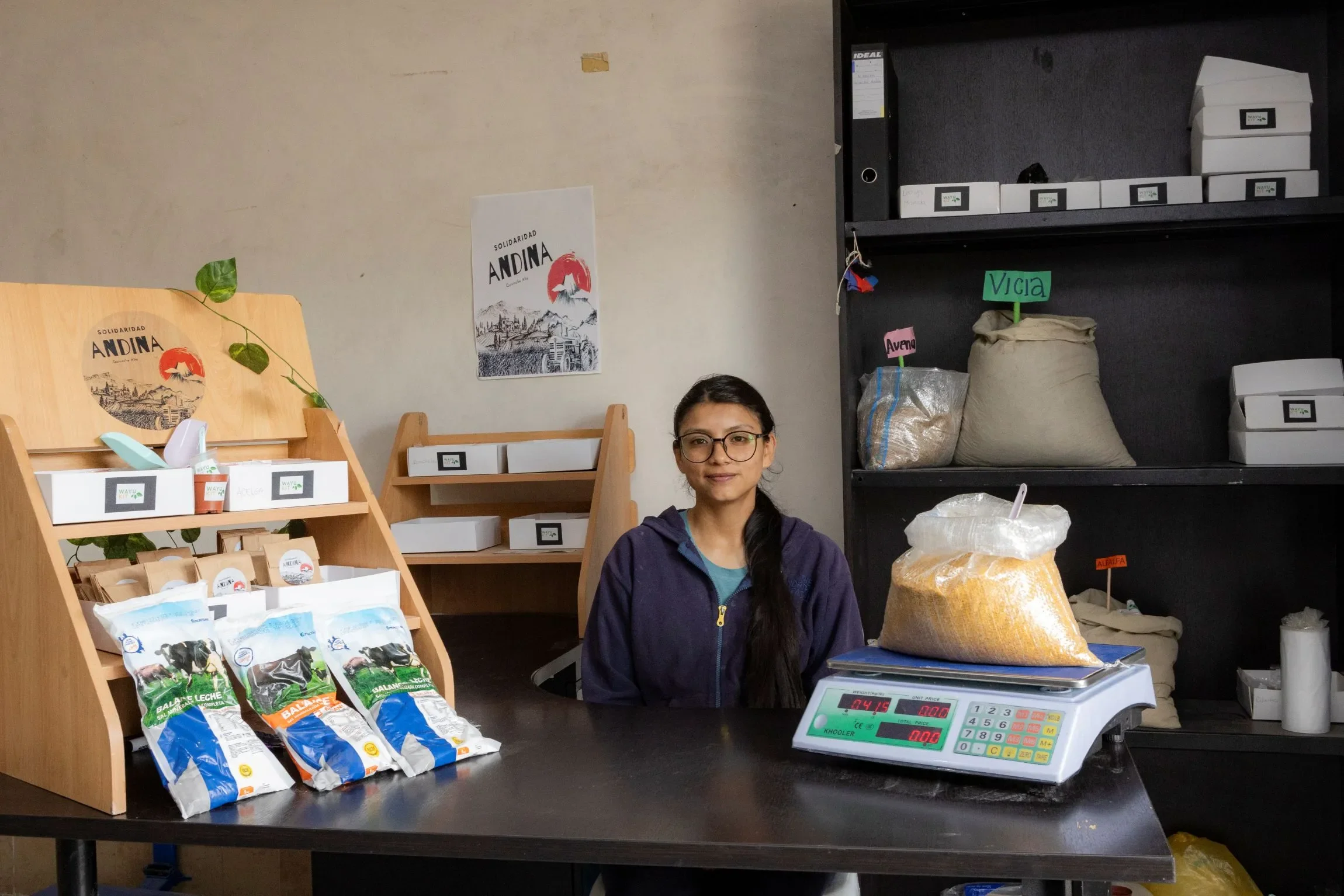 Yael, a Village Savings and Loan Association or VSLA member and young entrepreneur in Ecuador, sits behind the counter of her organic agricultural supply store, featuring wooden shelving, packaged goods, and a digital scale with a sack of grain.