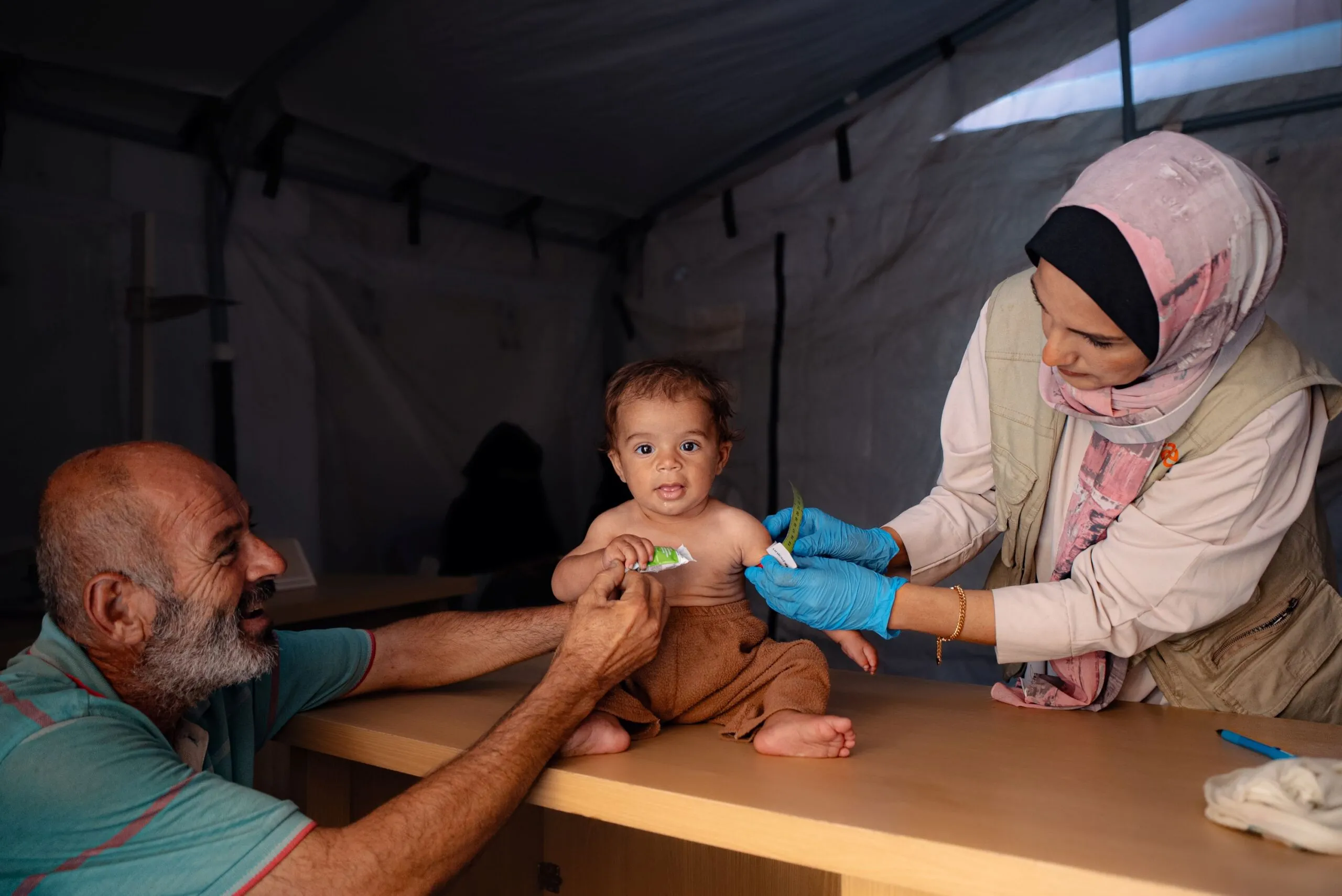 A healthcare worker in a clinic setting, wearing a hijab and blue medical gloves, examines a baby sitting on a wooden table. A man, likely the baby's father, kneels on the opposite side of the table, holding the baby's attention.