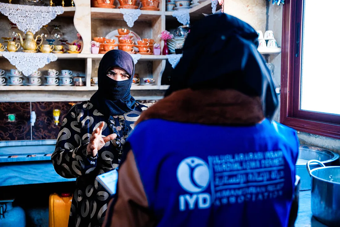 A Syrian woman wearing a headscarf and a patterned dress is talking to a person wearing a blue vest with the IYD humanitarian organization logo on it. They are in what looks like a small cafe or shop with shelves of teapots and cups.