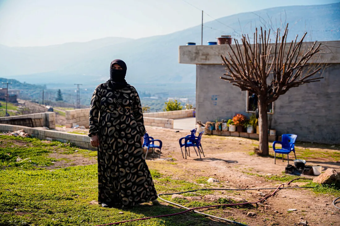 A woman wearing a patterned dress and a headscarf stands outdoors in a rural, grassy area. Behind her, there is a simple concrete building and a bare tree. In the background, there are hills under a clear blue sky.