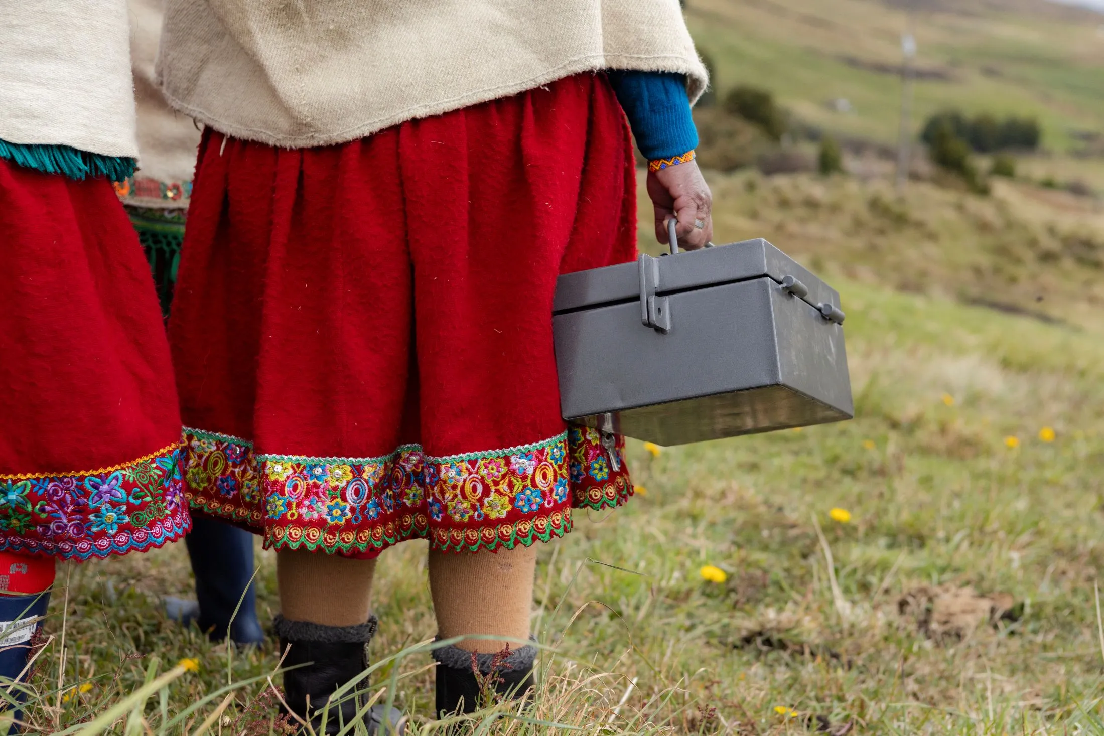 A VSLA or Village Savings and Loan Association member in Ecuador, wearing a traditional red skirt with colorful embroidered trim, stands in a grassy field holding the group's metal savings cash box.