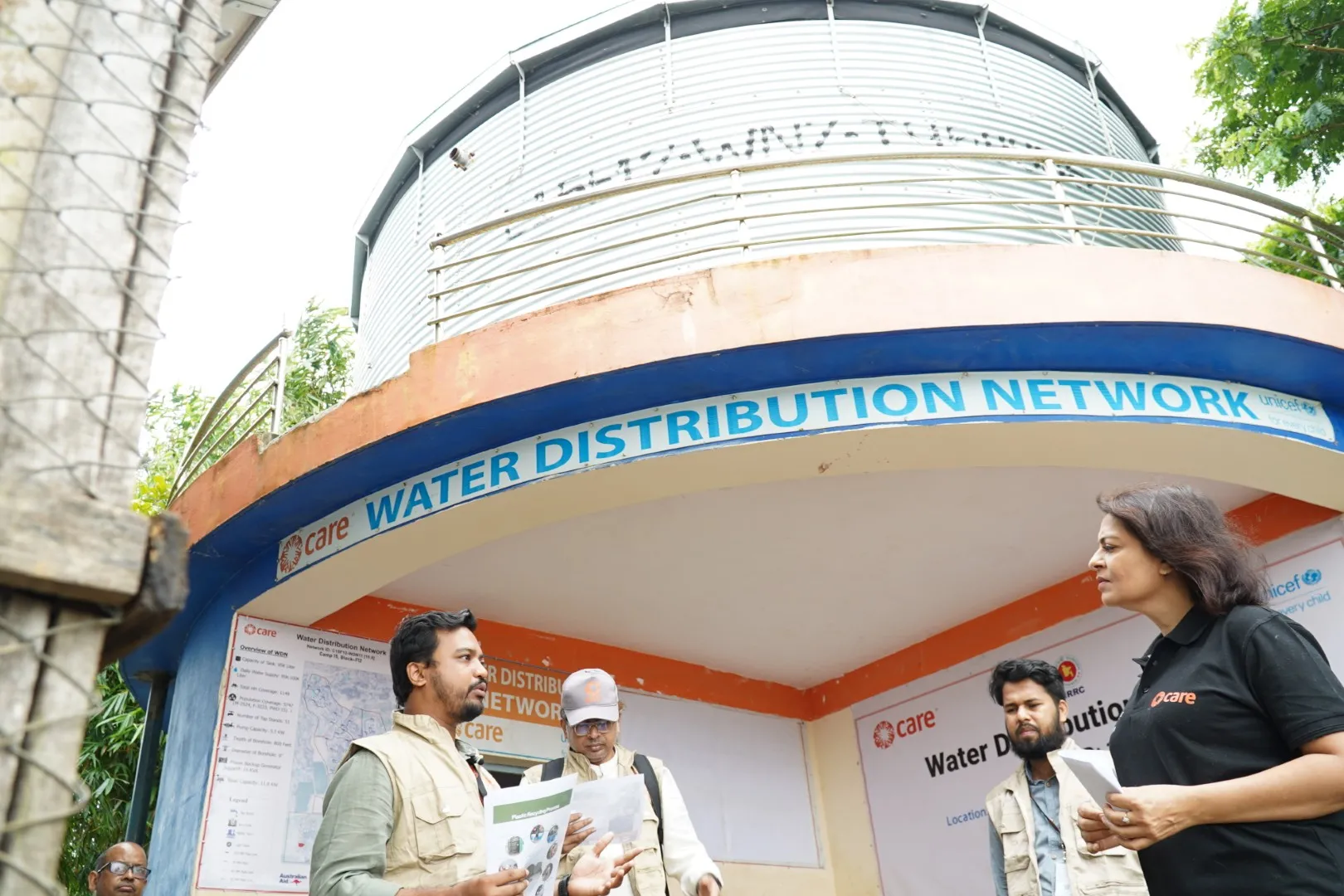 A group of humanitarian workers stands in front of a building and large water tank. Text on the building reads, 