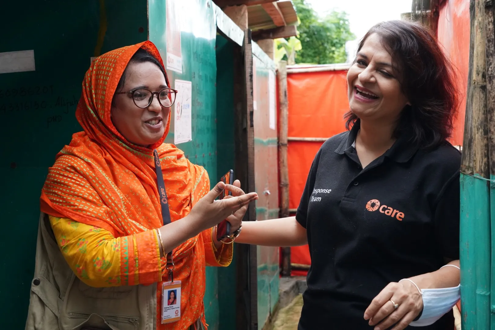 Two women, likely humanitarian workers, smile as they chat; one wears a black T-shirt with the CARE logo.