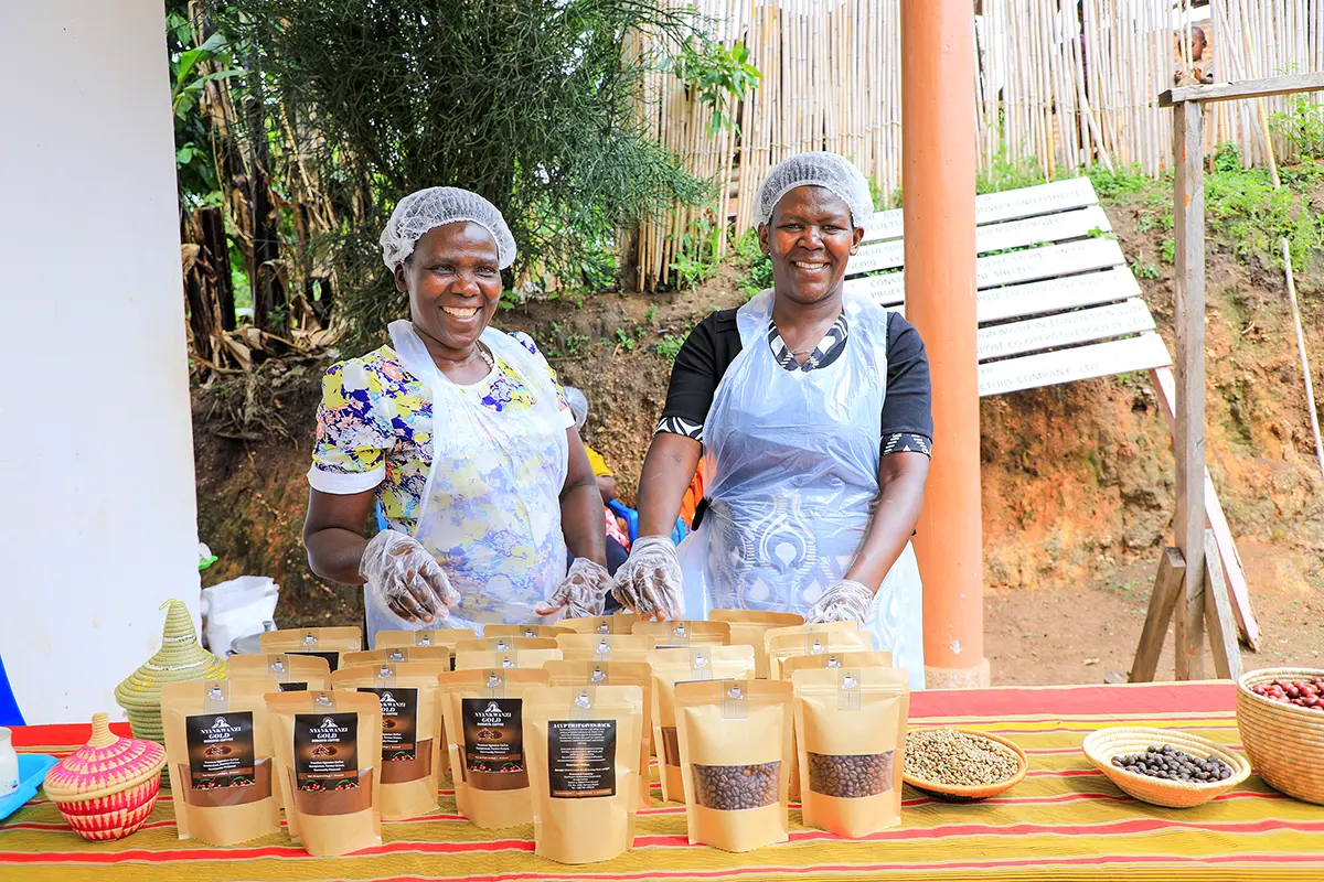 Two women wearing aprons and hair nets proudly stand behind their bagged coffee beans at a market counter.
