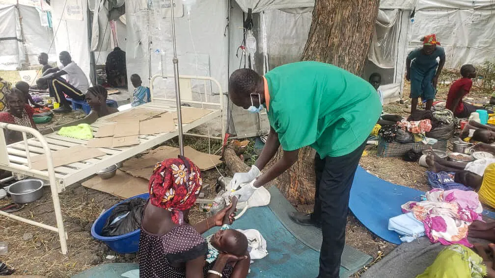 A medical professional treats a woman and child who sit on a tarp outside a health center.