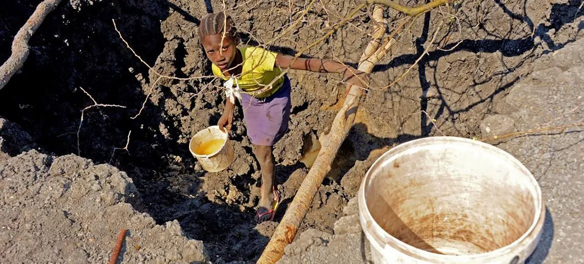 A young girl in a yellow shirt and purple skirt carefully balances over a narrow channel, holding a container to collect water. The ground is dry and cracked. She is looking at the camera, and the image is taken from above.