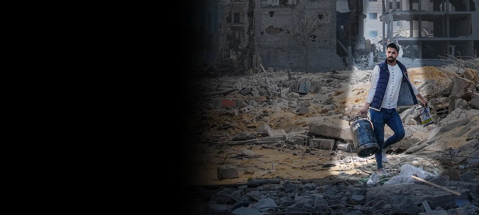 A Palestinian man carries a large can of water over rubble.