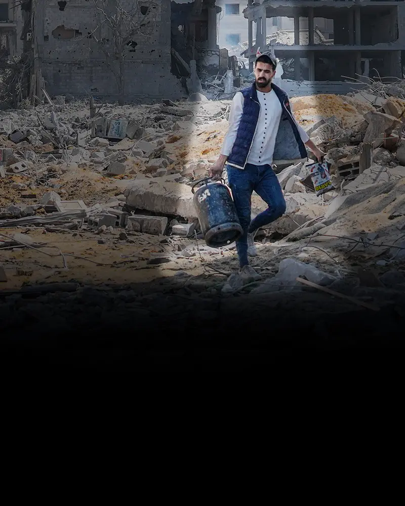 A Palestinian man carries a large can of water over rubble.