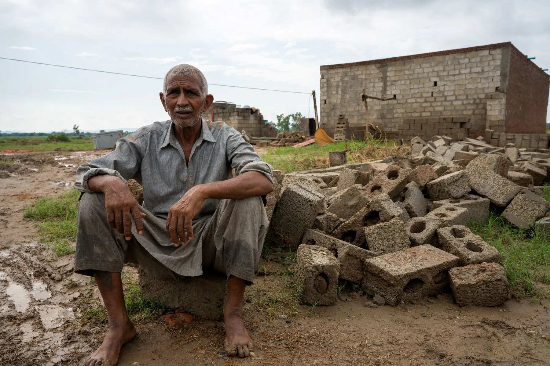 A weathered barefoot Pakistani man sits on broken cinder block in muddy, debris-strewn area with damaged building behind, under cloudy sky after disaster.