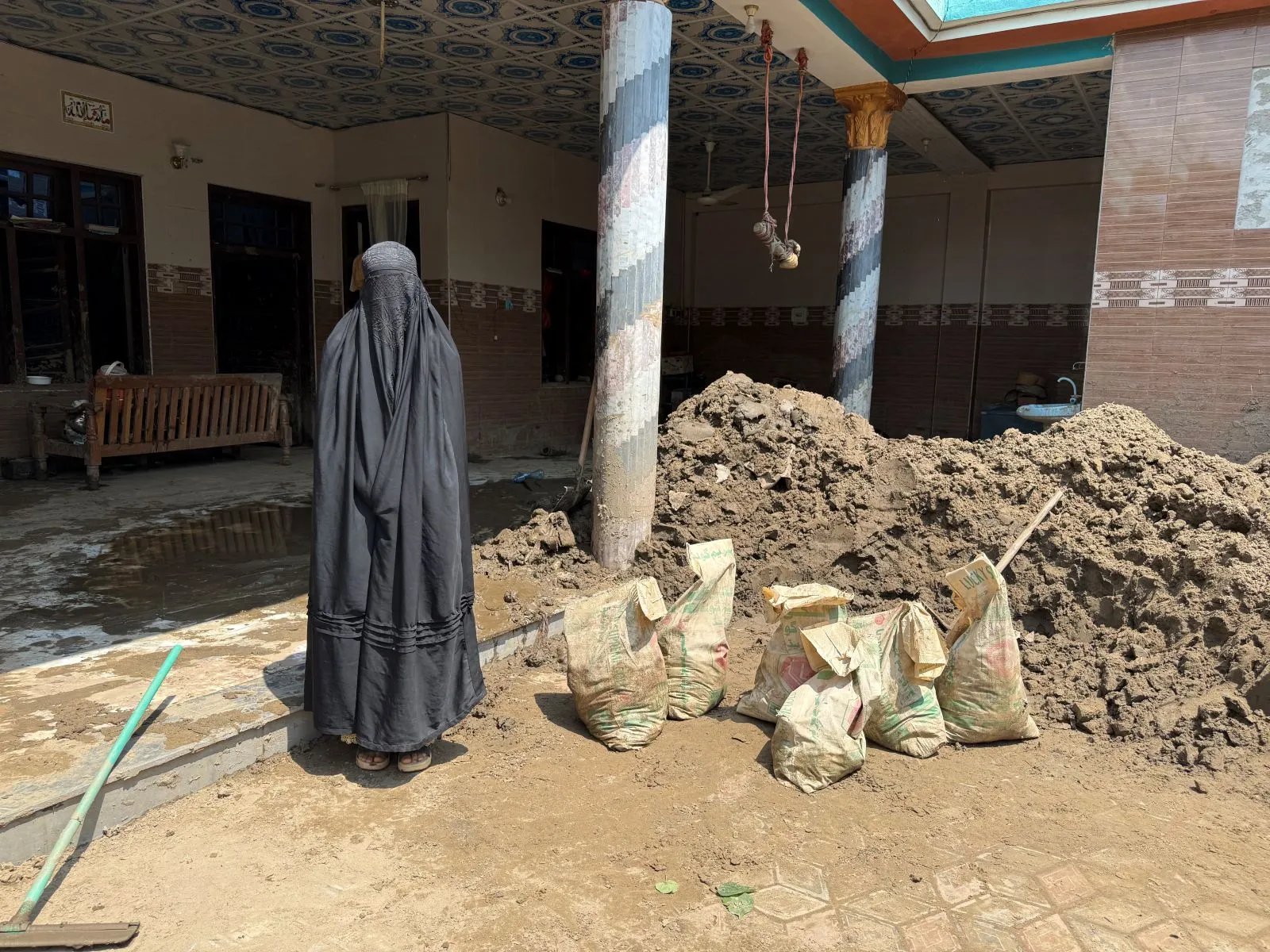 A Pakistani woman in a black burqa stands in a courtyard with muddy dirt and sacks. Behind her is a house with an ornate tiled roof and columns, and flood debris on the floor.