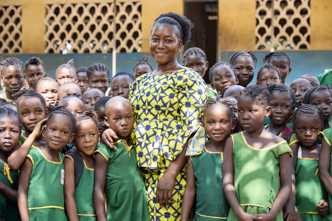 A woman in yellow stands surrounded by young children outside a school