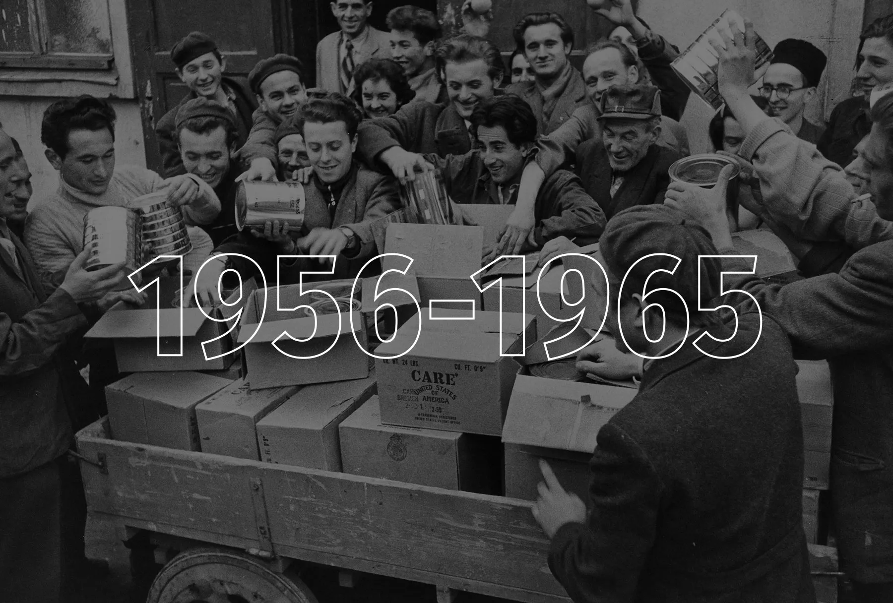 A large group of men surround a truck with CARE Packages and pass around the items inside.