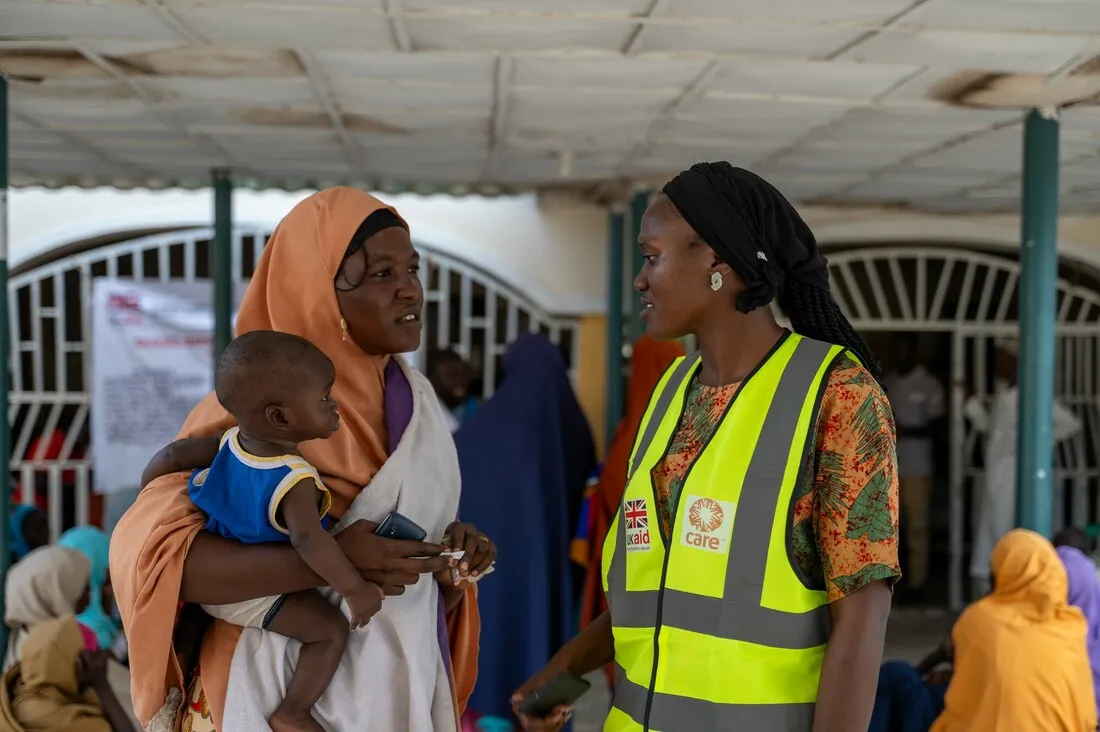 A woman wearing a neon yellow CARE vest talks with a woman holding a young child.