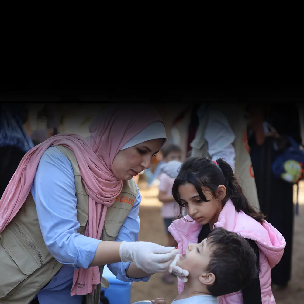 A woman wearing a pink headscarf and a khaki CARE vest administers the oral polio vaccine to a young boy.