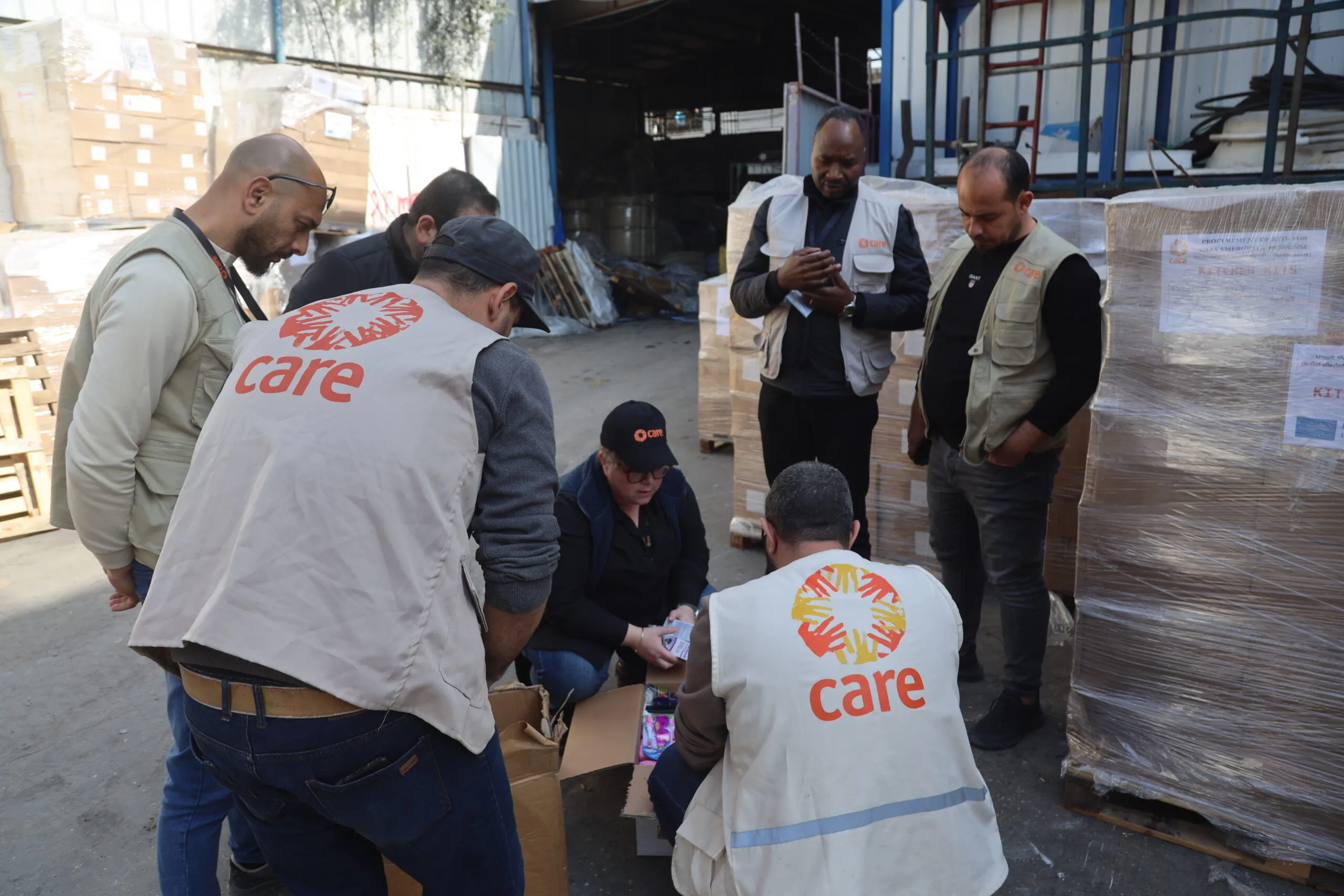 A group of people, several wearing CARE vests, crouch around packaged supplies in a warehouse in Gaza city.