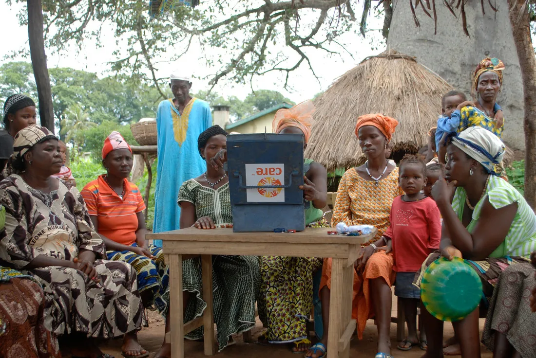 A group of several women sit around a table. On top of the table is an open gray metal box that says CARE.