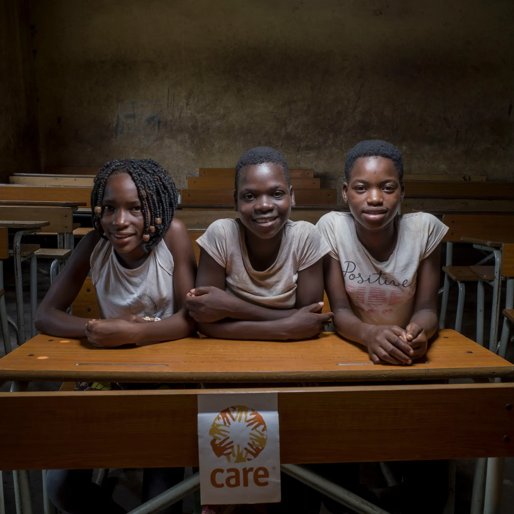 Three adolescent girls sit at a wooden desk in a classroom.