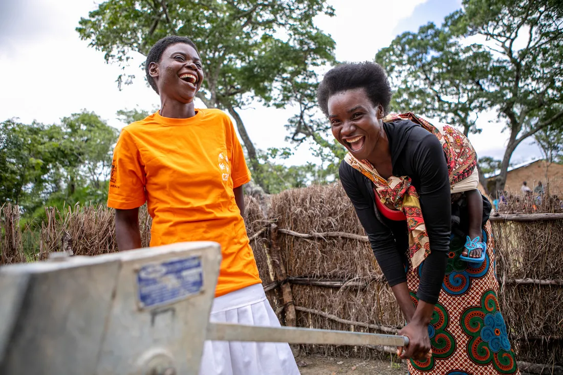 Two women, one wearing an orange CARE shirt, smile and laugh while using a water pump.