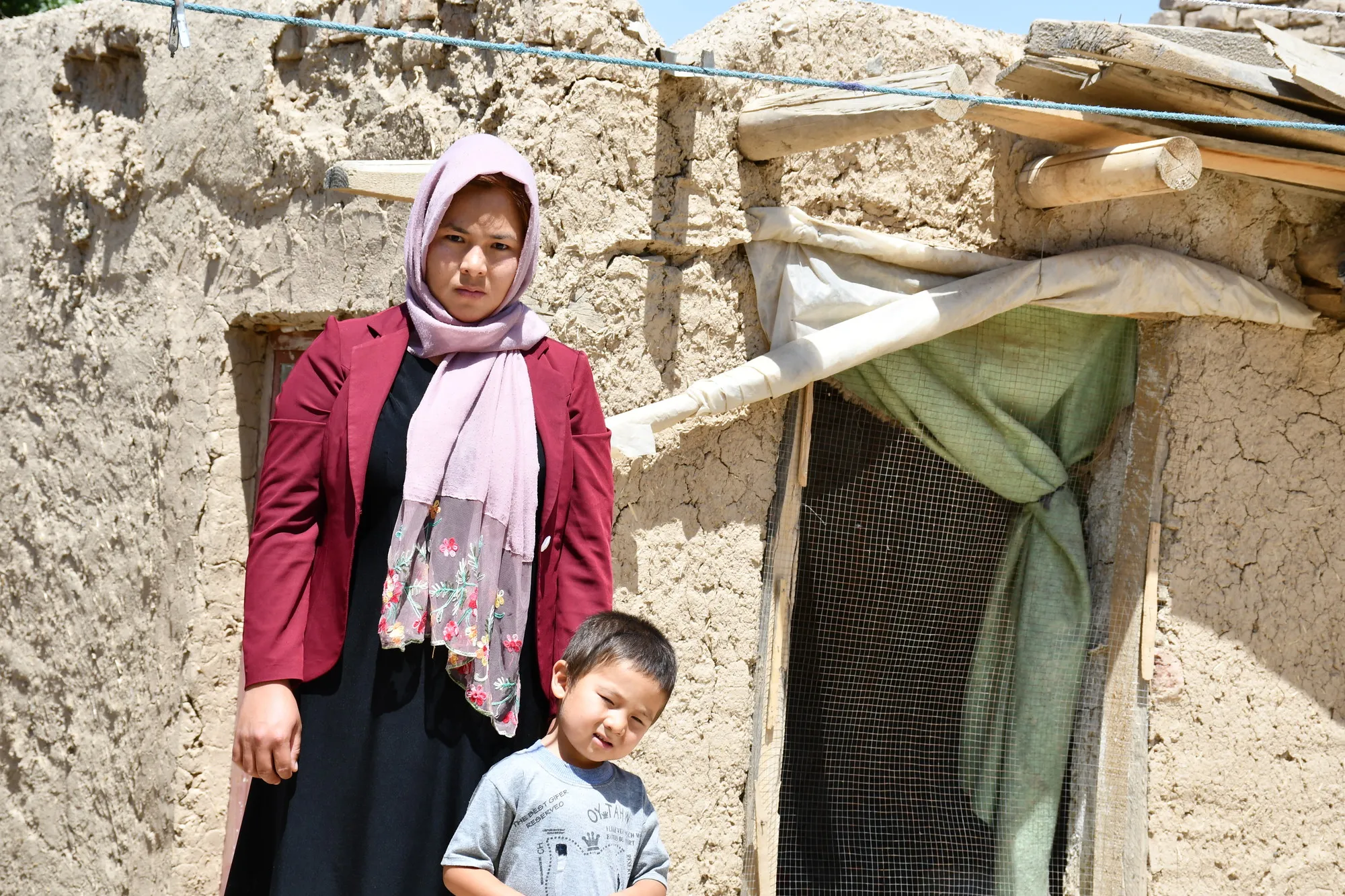 A woman and a small boy stand outside of a clay and wood home.