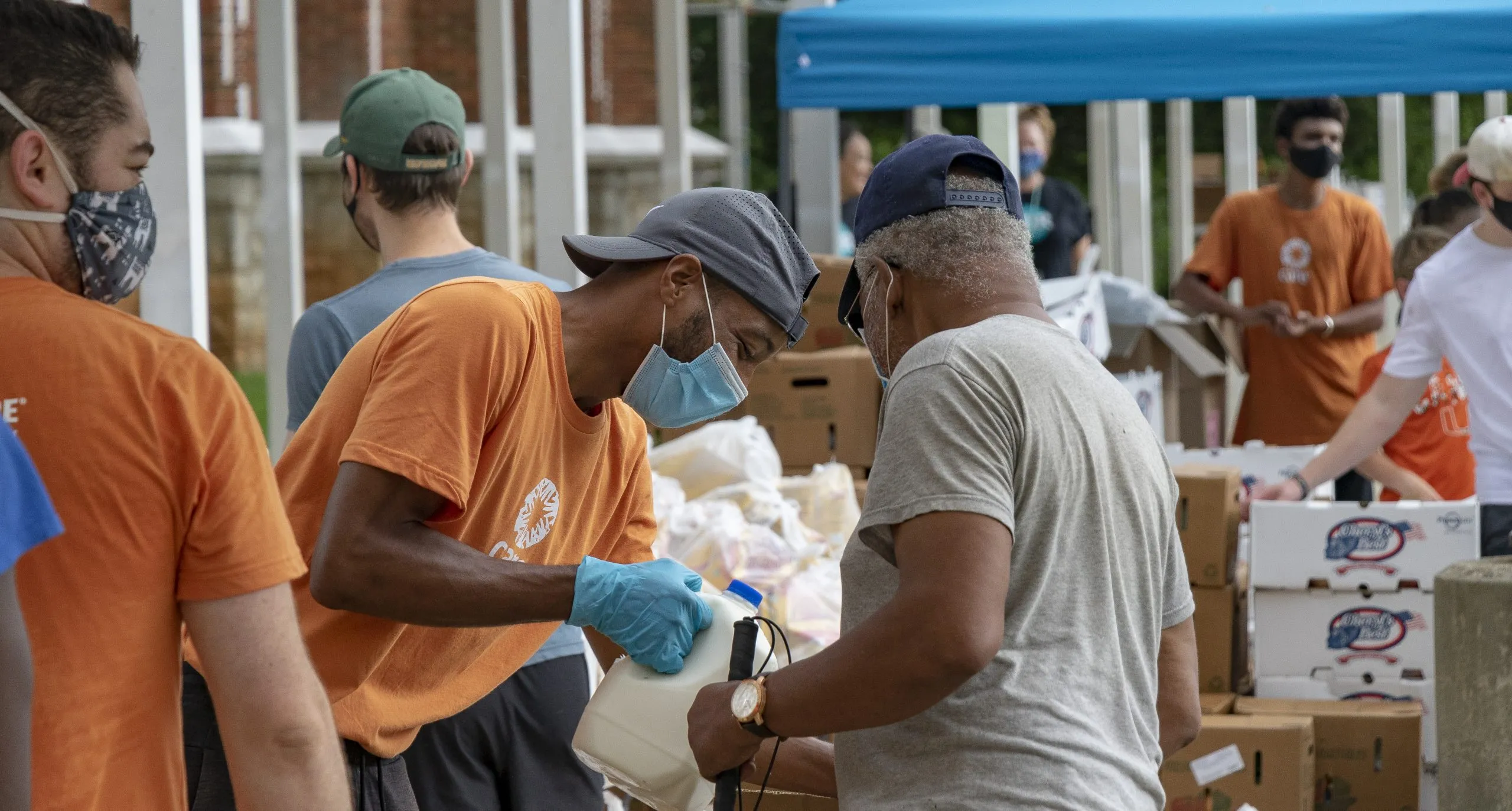 A man wearing an orange CARE t-shirt and PPE helps an older man with bagging groceries.