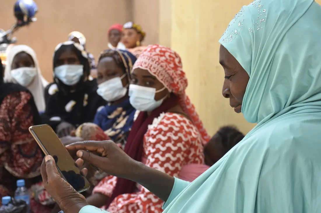 A woman wearing a light teal headscarf smiles while scrolling through a smartphone. Behind her are a group of women wearing facemasks and looking at her phone.