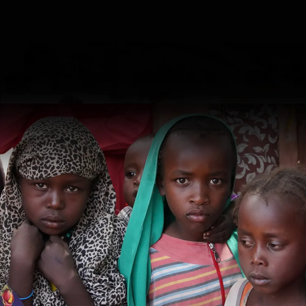 Three Sudanese children stand next to each other, one of them clutching fabric around their head.
