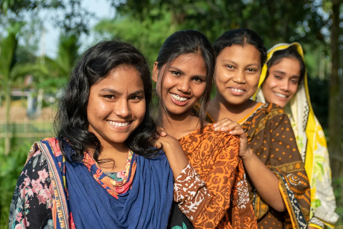 Four women stand together in a row with their hands on each others' shoulders.