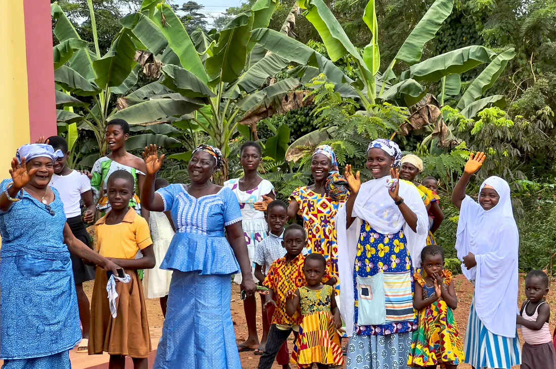 A group of Ghanian women and children, many wearing blue, white, and yellow, smile and wave.