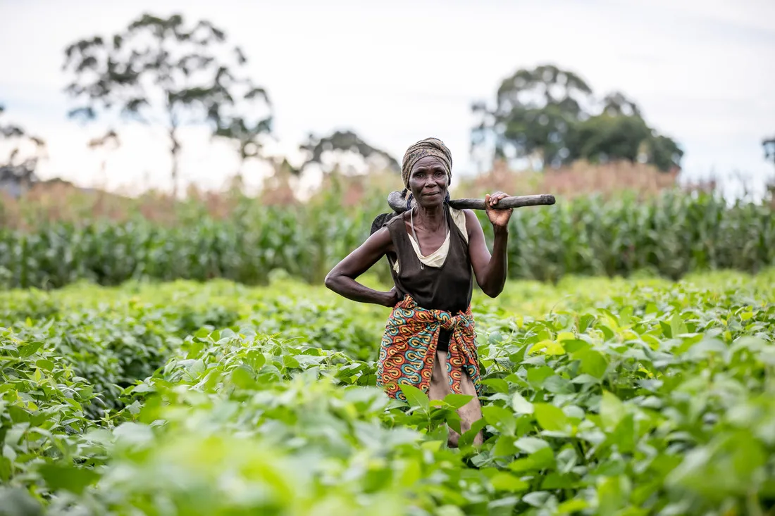 A woman stands in a field and rests a gardening hoe over her shoulder.